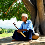 A wise-looking elderly East Timorese man, fully clothed in modest, traditional attire, is gently playing a Turina, a small, handcrafted stringed instrument. He is seated on a simple wooden stool under the sprawling branches of an ancient mango tree in a serene, sun-dappled traditional village. The scene captures a moment of cultural heritage and musical tradition, with soft, natural lighting. Perfect anatomy, correct proportions, natural pose, well-formed hands, proper finger count, natural body proportions. Safe for work, appropriate content, fully clothed, family-friendly.